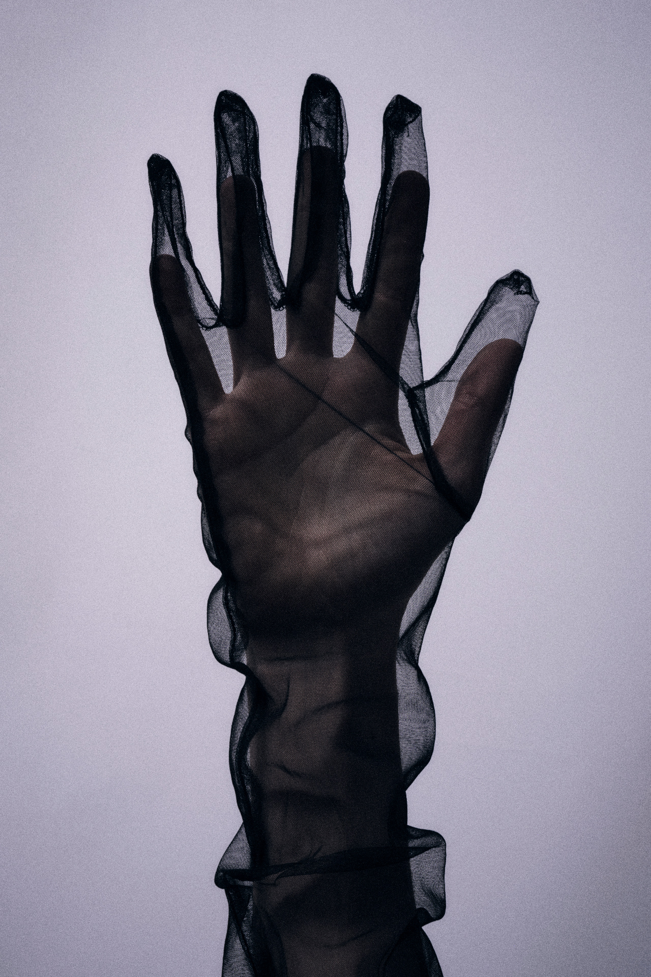 A raised hand in a sheer black glove against a pale wall