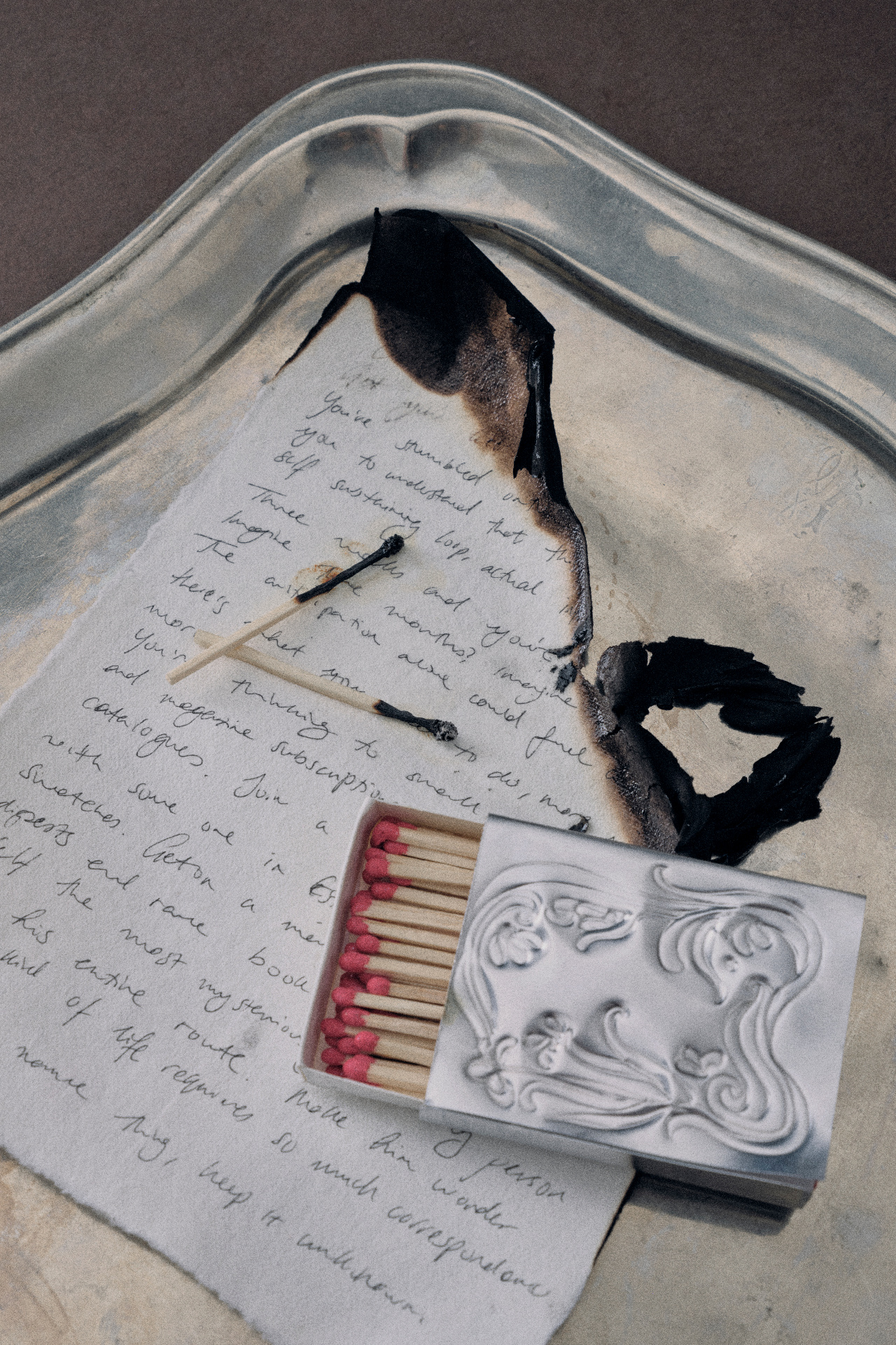 A partially burned handwritten letter on a silver tray, beside an open box of matches